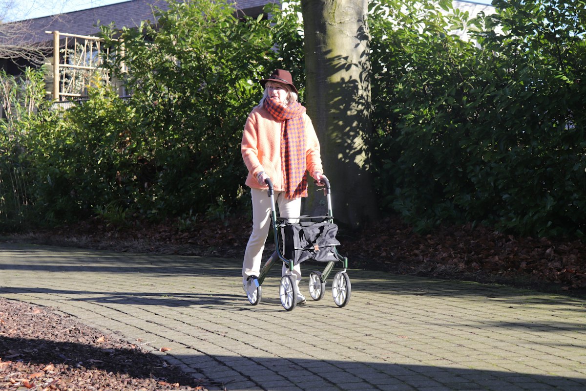 An older adult walking on a paved path with a rollator
